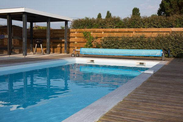 Swimmingpool mit Rollabdeckung, umgeben von einer Holzterrasse und grünen Büschen.